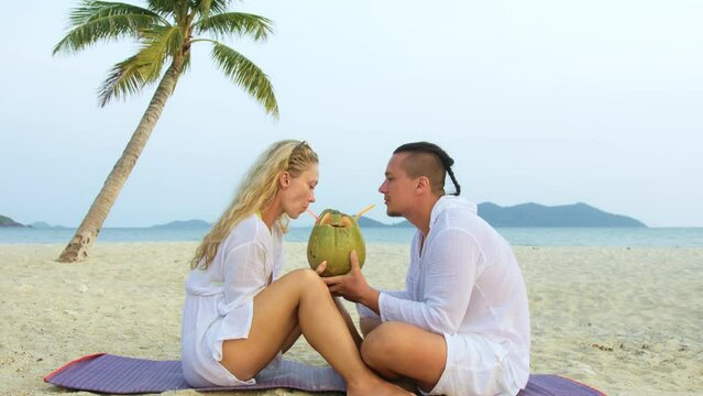 Carefree loving couple facing each other sits in tropical beach under palm tree and drink coconat cocktail, kissing and hugging. Concept honeymoon relationship romance beach tropical tourism