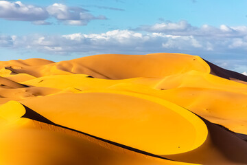 Fototapeta na wymiar Contrasted arc shaped and shadows of golden yellow and orange colored sand dunes with cloudy blue sky in the vast Sahara Desert of Algeria.