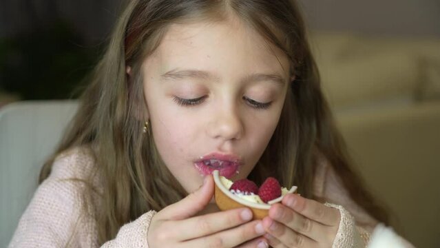 Child celebrates birthday with fresh raspberry tart. Adorable little girl eating fresh sweet raspberry cake indoors. Girl eating cake with cream and fruits.