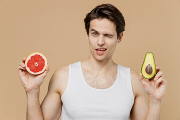Happy young man 20s perfect skin in undershirt hold half of avocado grapefruit choose wink isolated...