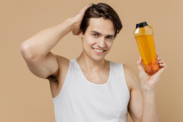 Attractive smiling happy young man 20s perfect skin in undershirt hold bottle of shampoo touch hair...