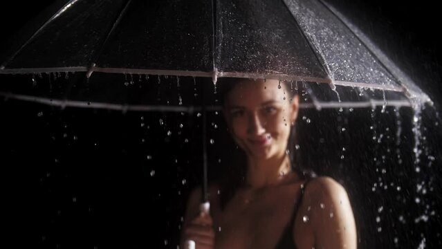 Young woman in black swimming suit standing under the rain and holding an umbrella - water drops dripping down from the edge