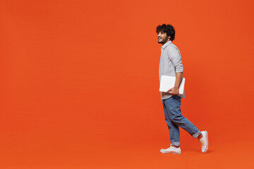 Naklejka premium Full size body length side profile view young bearded Indian man 20s years old wears blue shirt hold use work on laptop pc computer strolling pace isolated on plain orange background studio portrait.