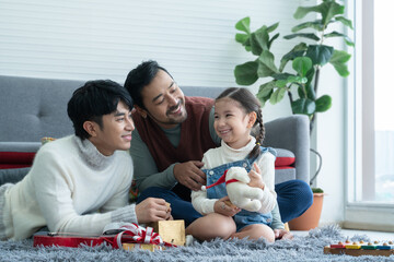 Asian young LGBTQ gay couple giving gift to little Caucasian adopted kid in living room at home....