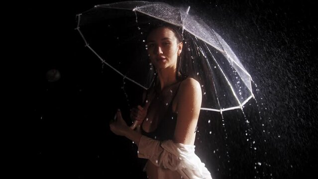 Young flirty woman in black swimming suit standing under the rain - holding and twirling an umbrella while looking at the camera