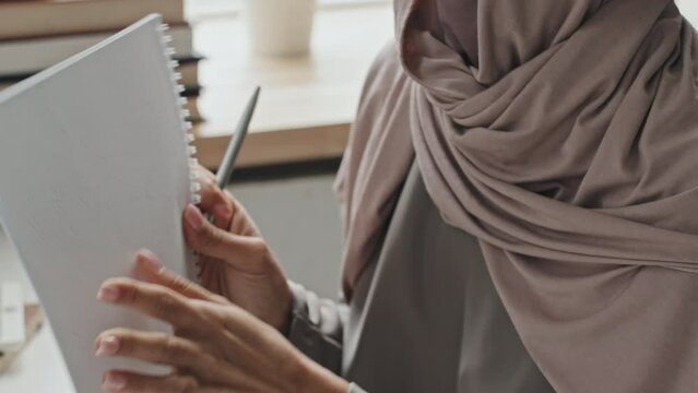 Close up shot of female teacher in hijab explaining task in workbook while giving online lesson