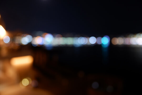 Cityscape with sea view in unfocused night lighting, blurred lights in white, blue, red, orange and yellow colors.