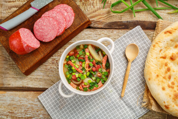 Jewish bean soup with smoked meats in a tureen on a gray napkin next to a sausage and a flatbread and a wooden spoon.