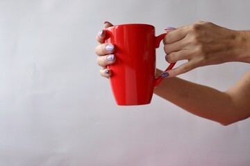 On a light background, female hands hold a large red cup. Gift for Valentine's Day. A woman holds out a cup to a man. Big cup of tea. Manicure color of the year 2022 - very peri.