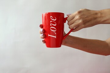 On a light background, female hands hold a large red cup with the inscription "Love".  Gift for Valentine's Day. A woman holds out a cup to a man. Big cup of tea. 