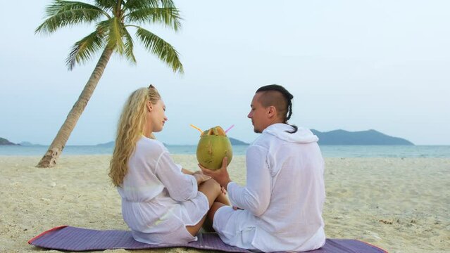 Carefree loving couple facing each other sits in tropical beach under palm tree and drink coconat cocktail, kissing and hugging. Concept honeymoon relationship romance beach tropical tourism