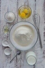 On a white wooden table are the ingredients for baking a dessert dish - flour, eggs, vegetable oil, sugar, salt, soda, milk or kefir. Photo for baking recipe.