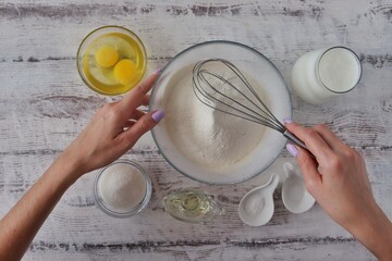 On a white wooden table are the ingredients for baking a dessert dish - flour, eggs, vegetable oil, sugar, salt, soda, milk or kefir. The woman is kneading the dough. Photo for baking recipe.