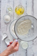 On a white wooden table are the ingredients for baking a dessert dish - flour, eggs, vegetable oil, sugar, salt, soda, milk or kefir. The woman is kneading the dough. Photo for baking recipe.