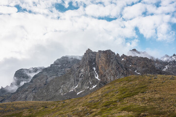 Dramatic landscape with sunlit hill and sharp pinnacle on high mountain range in low clouds. Beautiful alpine scenery with peaked mountain top in sunlight in low cloudy sky at changeable weather.