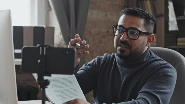 Middle eastern male tutor explaining task in workbook on camera of smartphone held by tripod while giving online lesson via video call
