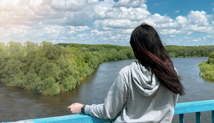 A dark haired caucasian girl stands with her back in a light gray jacket and looks towards the river and the forest