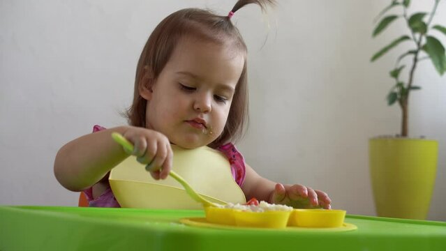 Close up of lovely baby girl eating have breakfast eggs vegetables sausage by herself at home. Adorable possitive happy toddler child eats with dirty face at table indoor. Food, nutrition healthy life
