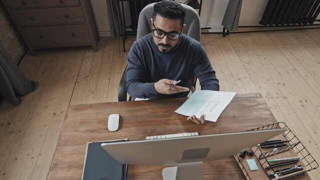 High angle shot of middle eastern male tutor showing text in workbook and talking on video call on computer while sitting at desk and giving online lesson