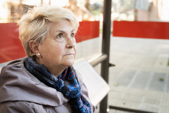 Portrait of senior blonde woman sitting on bus stop. Serious elder lady waiting for public transportation