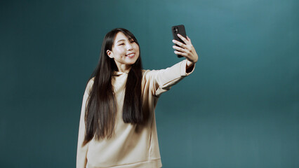 Pretty girl in beige hoodie looks into the screen of new mobile phone, makes selfie, shows her tongue