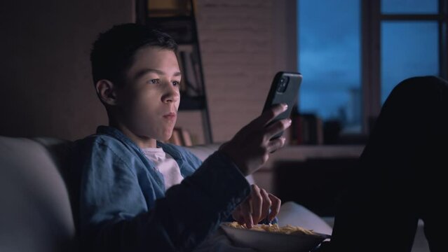 happy teen boy using phone at night at home and eating potato chips, chatting with friends, playing game indoors