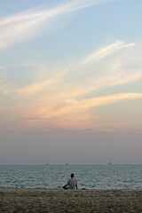 Fototapeta premium Back of a senior Asian man sitting on the sand at a beach looking at the ocean and the sunset sky, southeast Asia