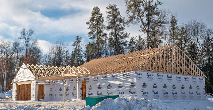 NISSWA, MN - 22 NOV 2021: Home construction site with Tyvek wrap, new wood roof trusses in place, and winter snow. Trees line the back of the building lot.