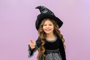A child in a witch's costume points his finger forward. A little beautiful girl in a magical Halloween masquerade.