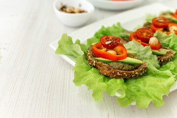 Vegan breakfast. Dietary nutrition. Sandwiches of grain bread with vegetables and nuts on a white plate on a white wooden table. Side view, close-up, space for text.