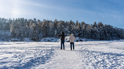 The trail passes through a frozen and snow-covered river. Footprints in the snow. Two people walk across the ice to the shore. Coniferous forest against the blue sky. Sunny  morning. Altai. Katun