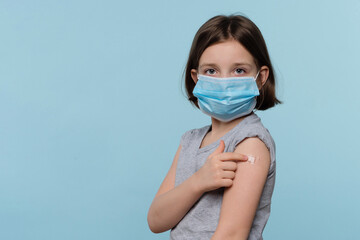 Kid girl in protective mask showing plaster patch on arm after vaccination against COVID-19...