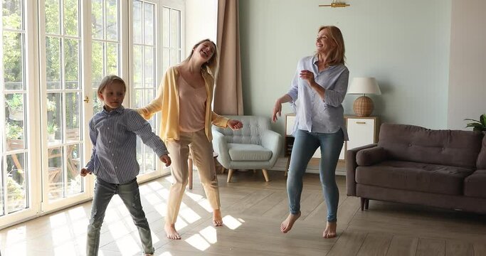 Overjoyed three generations of women dance barefoot on warm floor in living room relaxing on weekend. Happy energetic active 6s girl with young mom, 60s grandmother have fun home. Leisure concept