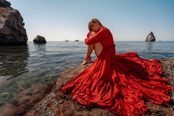 Beautiful sensual woman in a flying red dress and long hair, sitting on a rock above the beautiful...