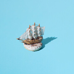 Small wooden home decoration pirate ship with white sails on donut with white glaze and colorful crumbs. Blue pastel ackground. Pattern. Minimal concept and design.