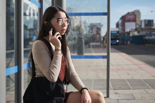 Young asian woman speaking on her smartphone while waiting for the bus at bus stop.