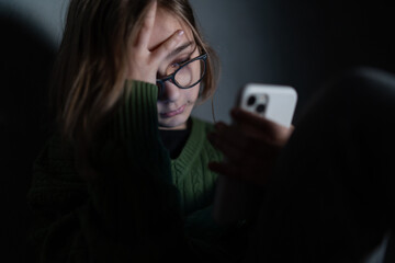 Sad little girl, alone in darkness, sitting and using smartphone.