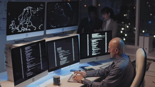 Side view of bold mature Caucasian man wearing formal shirt and eyeglasses, sitting at desk in office at night, coding, two blurred colleagues talking on background
