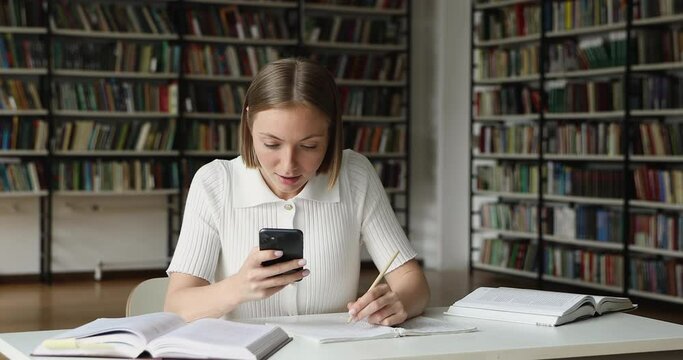 Confident young female pupil student use phone in self studying search data at web sources. Teenage lady learner make notes from smartphone screen using free access to wifi internet at public library