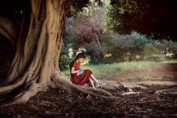 Cute european kid girl in a dress sits near a big tree in the park. A child writes poetry on large tree roots, a girl makes notes - obrazy, fototapety, plakaty