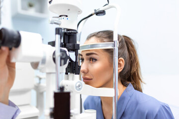Attentive optometrist examining female patient on slit lamp in ophthalmology clinic. Young beautiful woman is diagnosed with eye pressure on special ophthalmological equipment. - obrazy, fototapety, plakaty