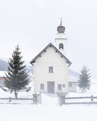 Fototapeta na wymiar Small church of the Immaculate of Viera in Livigno, Italy in severe snow storm. Snow blizzard and mountain in background, minimalist composition