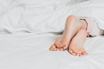 Childs legs are covered with white bed linen. The warmth of home comfort.