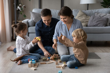 Affectionate millennial couple parents playing toys with adorable children son daughter, having fun enjoying playtime activity sitting together on floor carpet in living room, family relations concept
