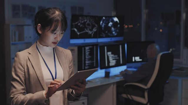 Waist-up of young Asian businesswoman standing in corporate office at night, using tablet computer, blurred programmer coding on background