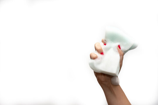 Mano de mujer joven con u&ntilde;as rojas apretando con los dedos una esponja para limpiar con mucha espuma blanca sobre un fondo blanco