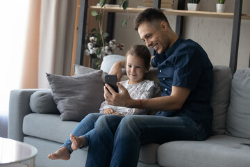 Joyful bonding millennial father and small cute kid daughter looking at smartphone screen, watching funny photo video content in social network, playing games or shopping online, purchasing goods.