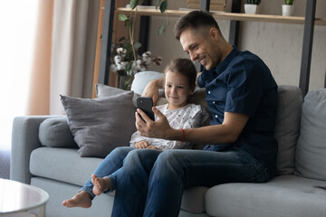 Obraz premium Joyful bonding millennial father and small cute kid daughter looking at smartphone screen, watching funny photo video content in social network, playing games or shopping online, purchasing goods.