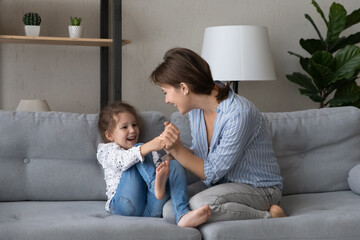Loving caring young mother tickling cute small kid daughter, having fun together in modern living...
