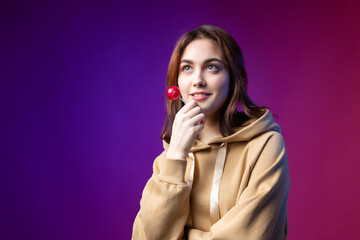 Portrait of a woman in a hoodie, girls licking a red round lollipop with beautiful makeup on a purple trendy background in the studio.